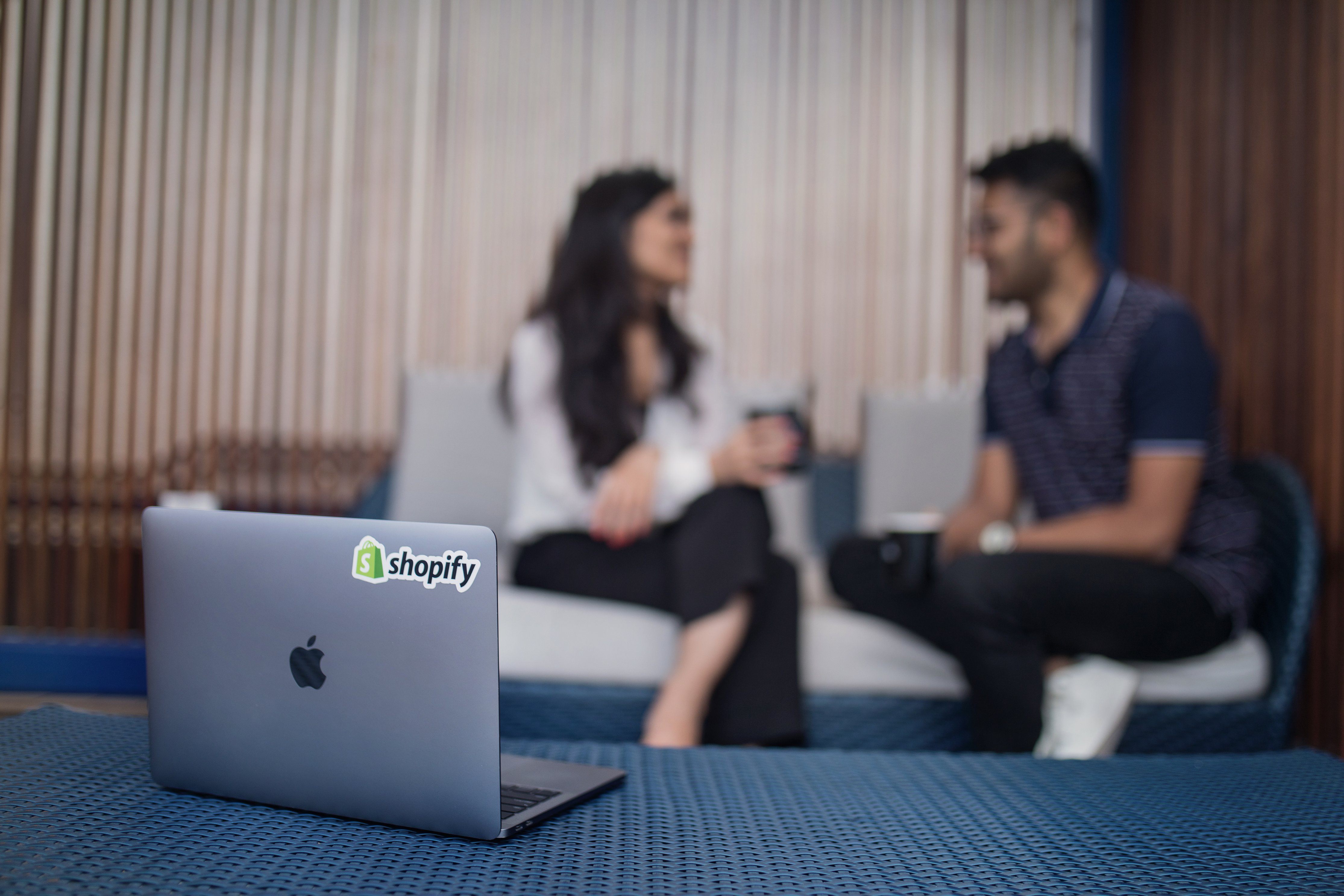 Two people having a conversation behind a laptop with a sticker with a Shopify logo 